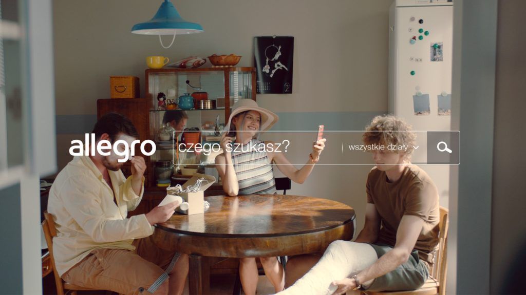 Allegro. Kadr z filmu „Czego szukasz” rodzina siedzi przy okrągłym stole w pokoju dziewczyna mierzy kapelusz przeglądając się w telefonie.